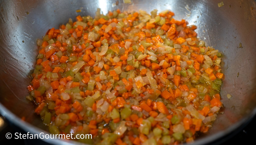 Finely diced carrots, celery, and onion sautéed in a large pan, creating a colorful mirepoix.