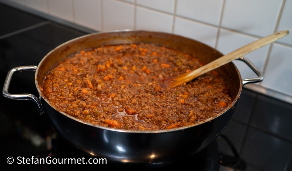 A large pot of Bolognese sauce with ground meat and diced vegetables simmering on a stovetop, with a wooden spoon stirring the mixture.