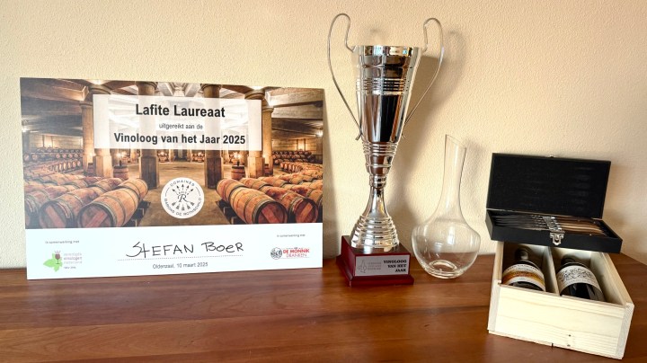A trophy and award certificate for 'Vinoloog van het Jaar 2025' displayed on a wooden table, alongside a glass decanter and a wooden box containing wine bottles.
