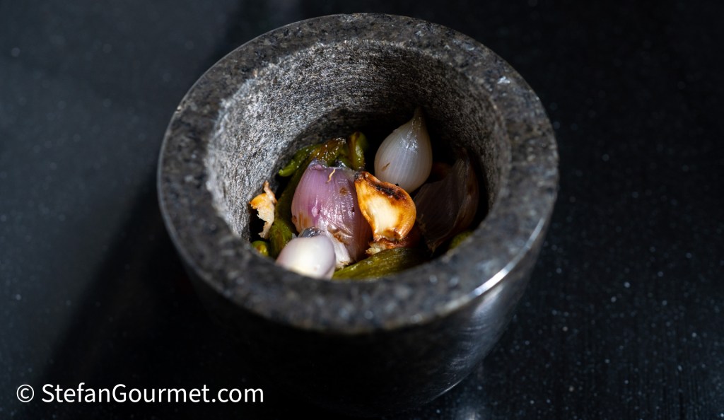 A stone mortar filled with roasted green chillies, shallots, and garlic, ready for making Nam Phrik Num, a Thai chili dip.