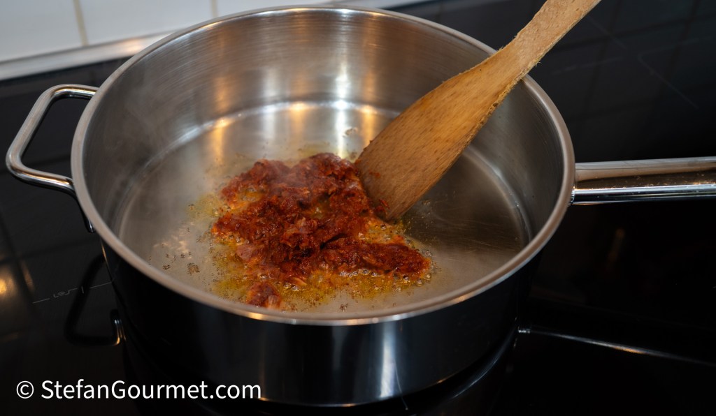 Cooking chili paste in a stainless steel pot on a stovetop, with a wooden spoon stirring the mixture.