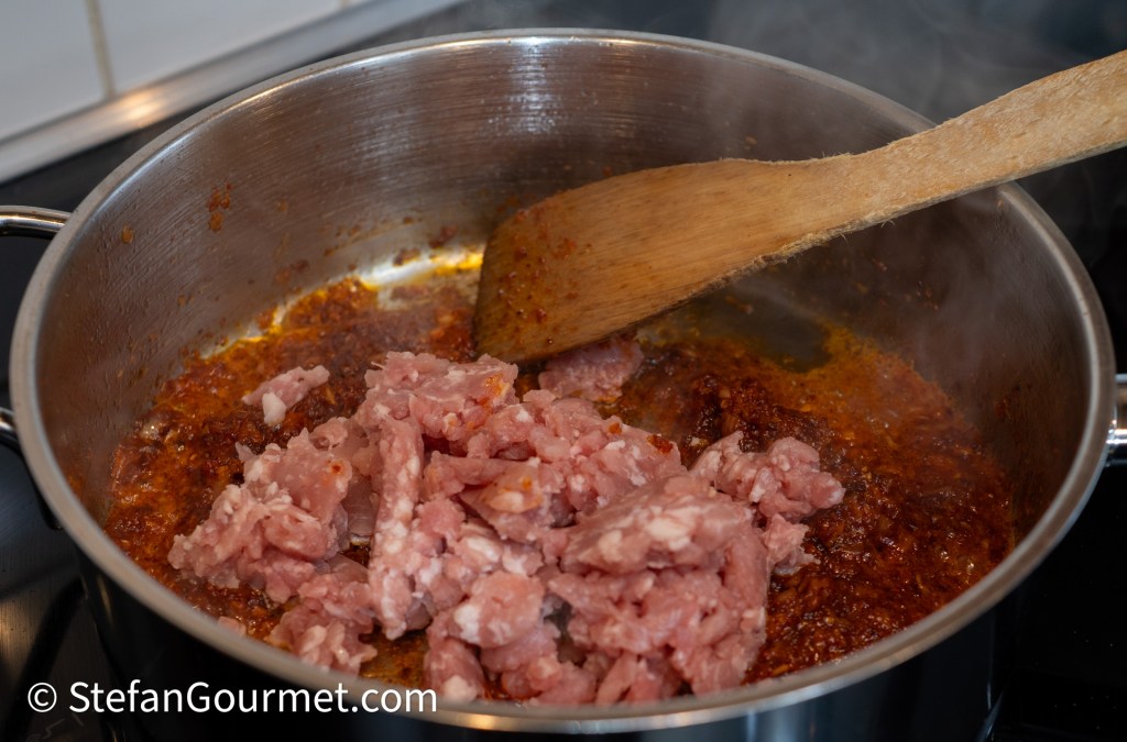 Ground pork being added to a simmering chili paste in a stainless steel pot, with a wooden spoon in action.