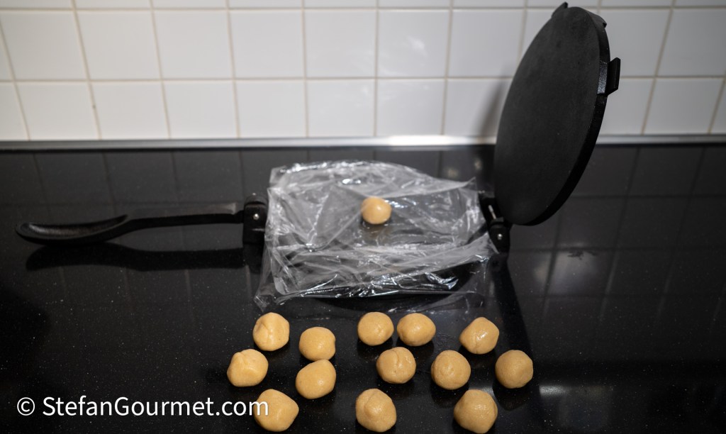 A tabletop with a tortilla press and several small balls of dough made from almond meal, resting on a sheet of cling film.
