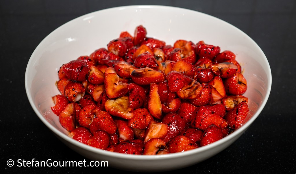 A white bowl filled with fresh, halved strawberries, some glistening with a honey and balsamic glaze.