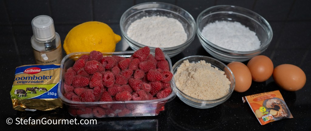 Ingredients for raspberry and almond cake, including raspberries, lemon, flour, almond meal, butter, sugar, eggs, and baking powder arranged on a countertop.