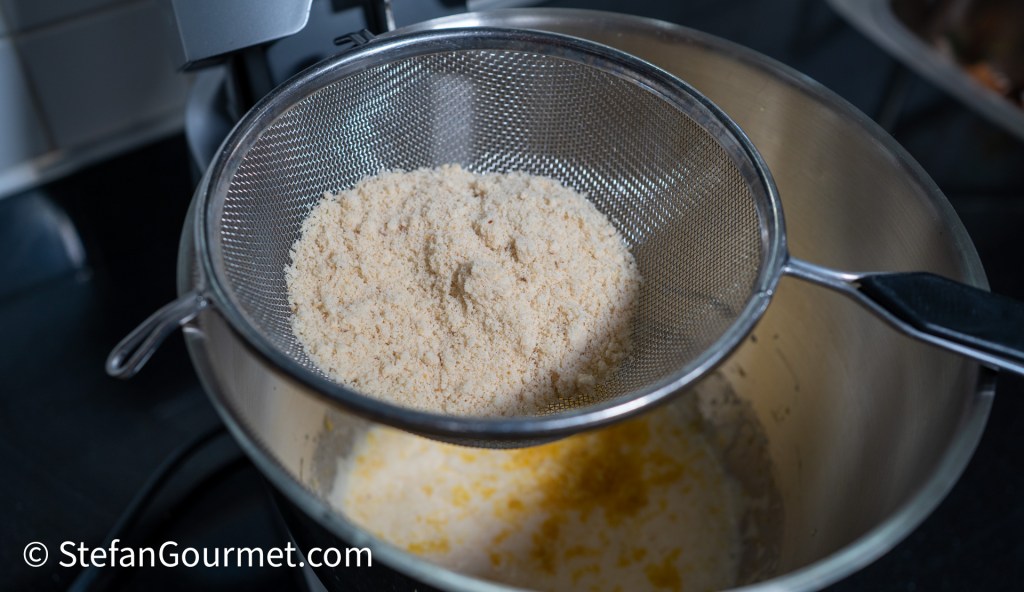 A close-up of almond meal being sifted into a mixing bowl, with lemon zest and a liquid mixture visible at the bottom.