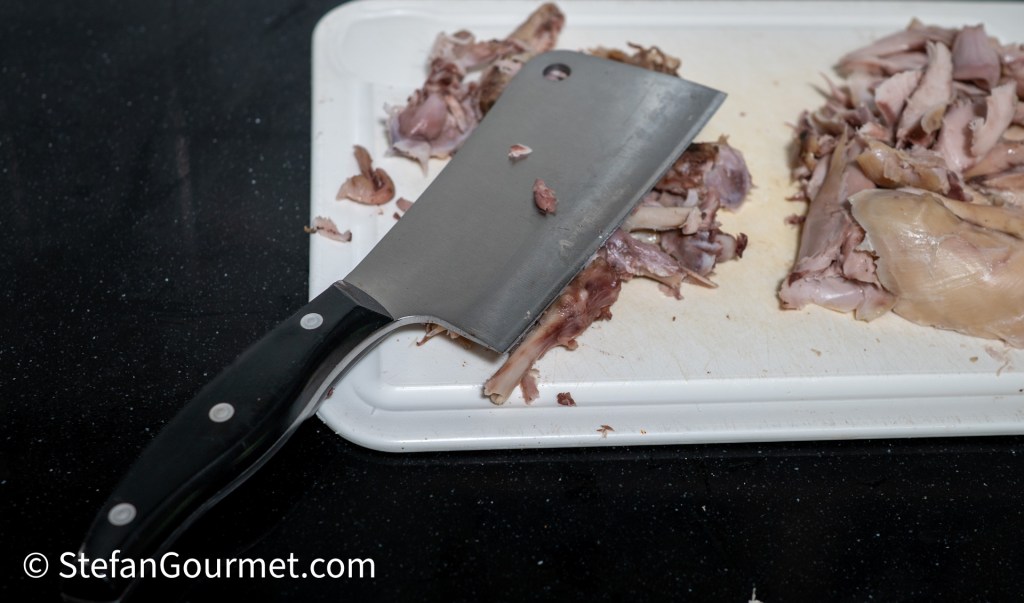 A cleaver resting on a cutting board with pieces of rabbit meat chopped into small bits.