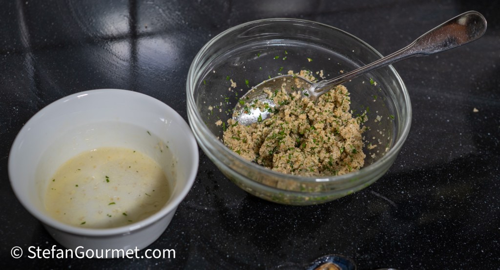 A glass bowl containing a breadcrumb mixture with parsley, alongside a small white bowl with a reserved mussel liquid, set on a dark countertop.