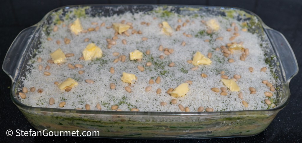 A glass baking dish filled with lasagna, topped with grated cheese, butter pieces, and pine nuts.