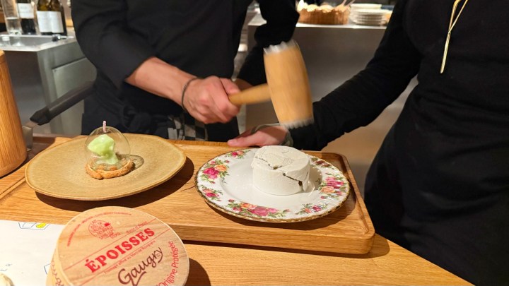 A chef using a wooden mallet to crack a dessert on a floral plate, with a green dessert in a glass dome on a wooden tray.