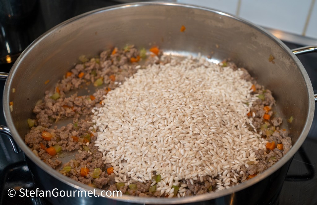 A large pan with cooked Italian sausage, diced vegetables, and uncooked risotto rice piled on top, ready to be stirred together.