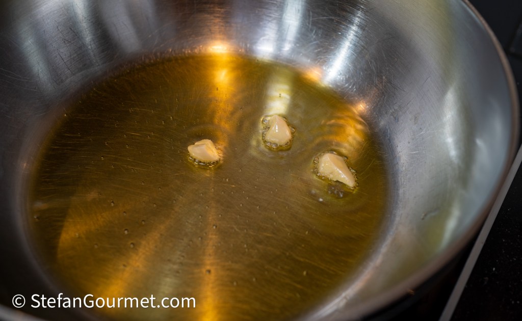 A stainless steel frying pan with olive oil heating up, featuring pieces of garlic melting in the oil.