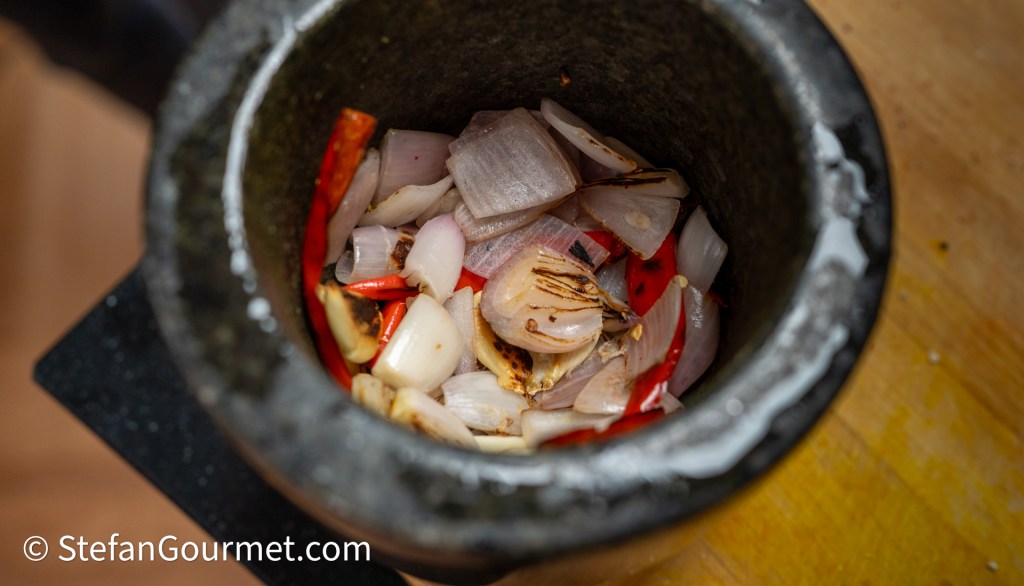 A stone mortar filled with sliced onions, red chili peppers, and garlic, ready for grinding.