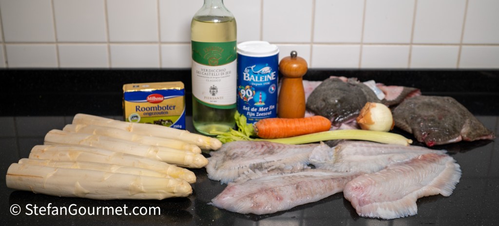 An assortment of cooking ingredients including white asparagus, various types of fish fillets, a bottle of white wine, a box of butter, salt, a carrot, celery, an onion, and a pepper grinder placed on a black countertop.