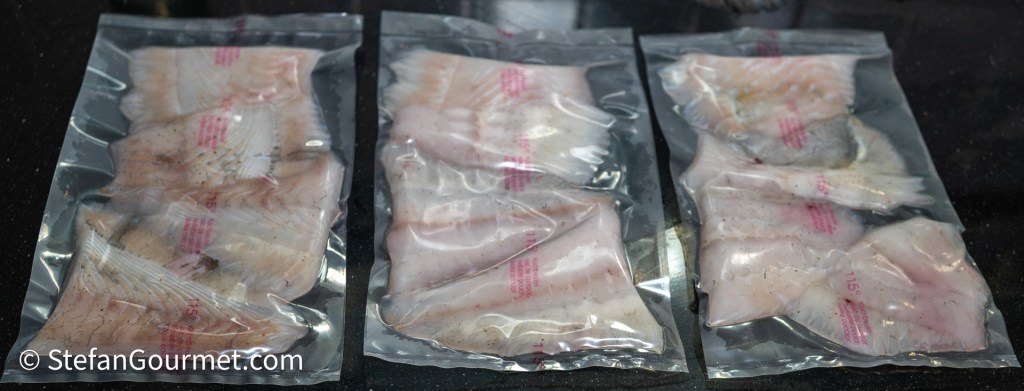 Three vacuum-sealed packages containing fish fillets arranged side by side on a dark surface.