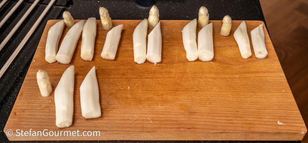 A wooden cutting board with various sizes and shapes of cut white asparagus spears arranged neatly.