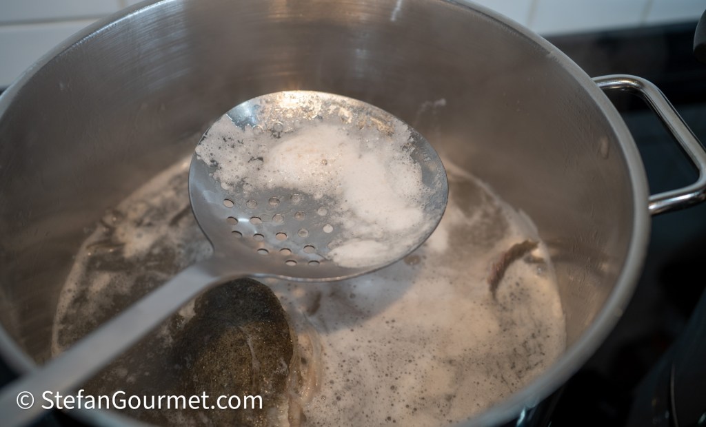 A metal slotted spoon lifting foam from boiling water in a stainless steel pot.
