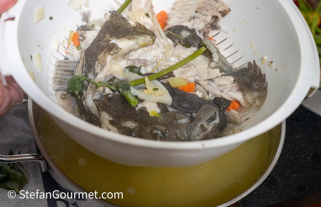 A hand holding a colander filled with fish bones and vegetables, including chopped carrots and green onions, above a pot of broth.