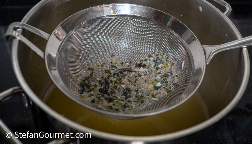 A stainless steel sieve resting over a pot, containing strained herbs and solids from a broth, with yellow liquid visible in the pot below.
