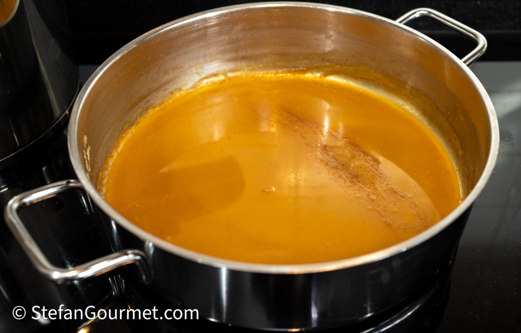 A stainless steel pot filled with a smooth, golden-brown sauce or broth, reflecting light on its surface.