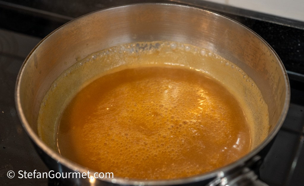 A stainless steel pot filled with simmering golden-brown liquid on a stove, with bubbles forming on the surface.