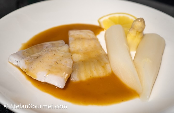 A plate featuring two fillets of fish drizzled with a brown sauce, accompanied by white asparagus, a slice of lemon, and a small piece of lemon zest.