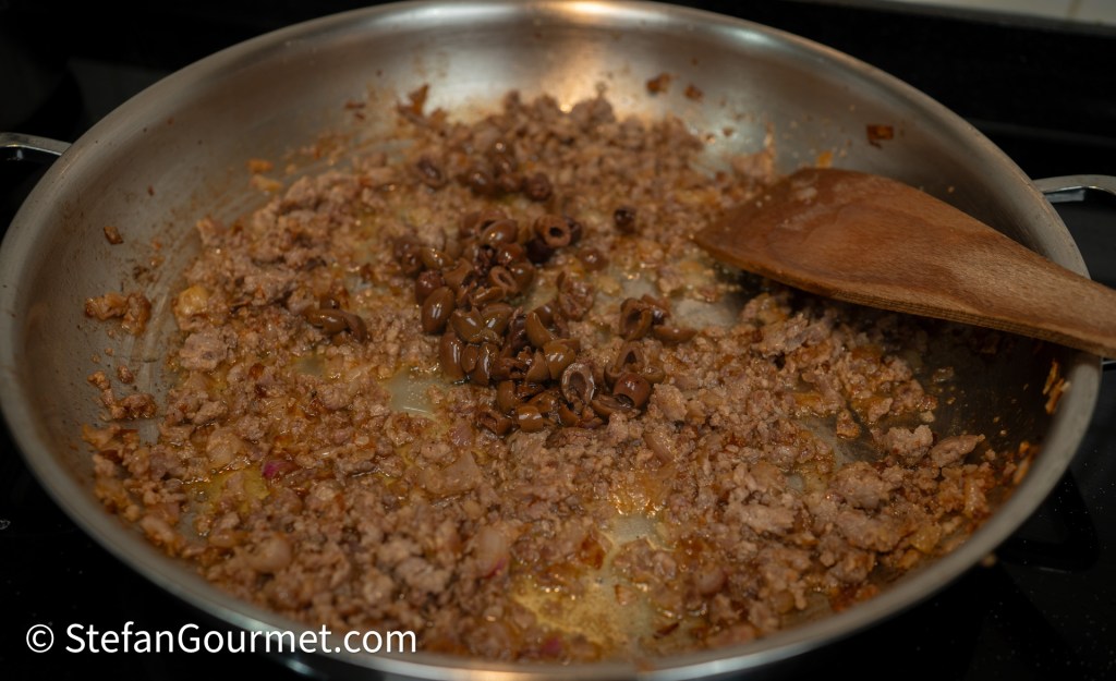 Cooked ground meat mixed with sliced olives in a frying pan, with a wooden spoon resting on the side.