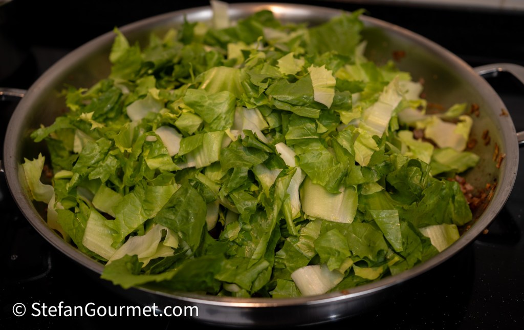 A close-up of a skillet filled with large, chopped romaine lettuce leaves, resting on a stovetop.