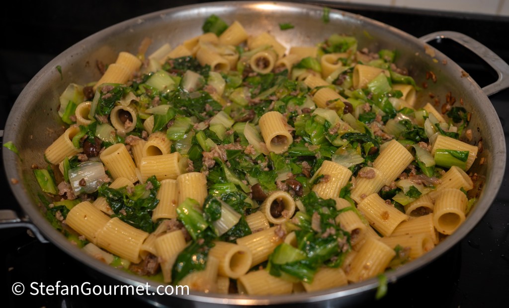 A skillet filled with cooked rigatoni pasta mixed with chopped greens and ground meat, garnished with various vegetables.