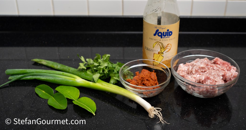 An assortment of ingredients for cooking, including green onions, fresh cilantro, kaffir lime leaves, a jar of premium fish sauce, a bowl of ground meat, and a small dish of red curry paste, all arranged on a black countertop.