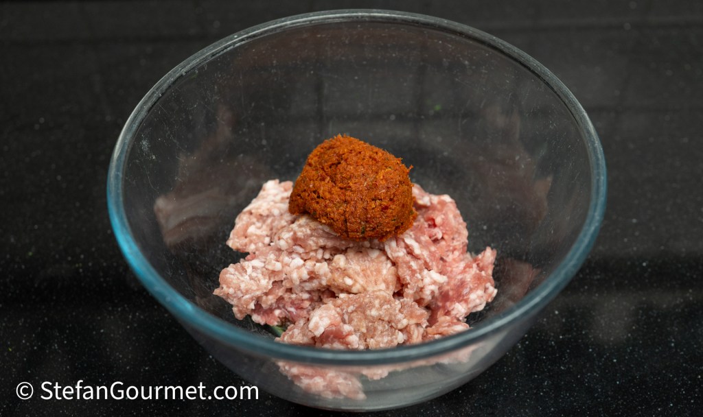 A glass mixing bowl containing raw ground meat and a ball of seasoning paste, placed on a dark countertop.