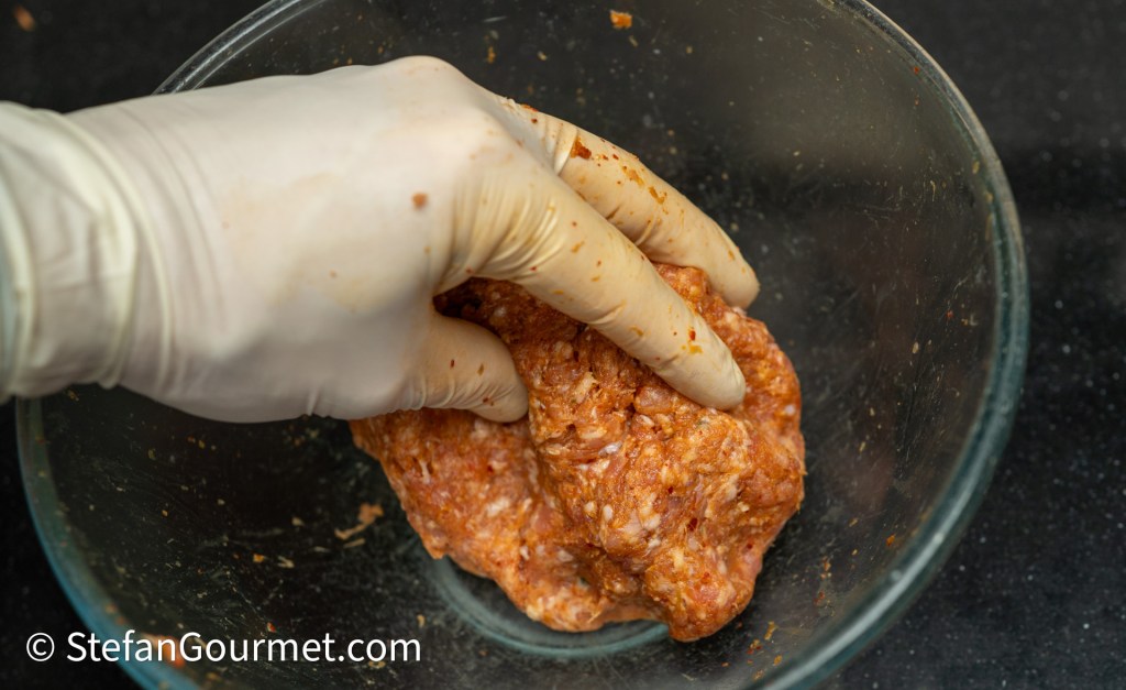 A gloved hand mixing a seasoned meat mixture in a glass bowl on a dark surface.