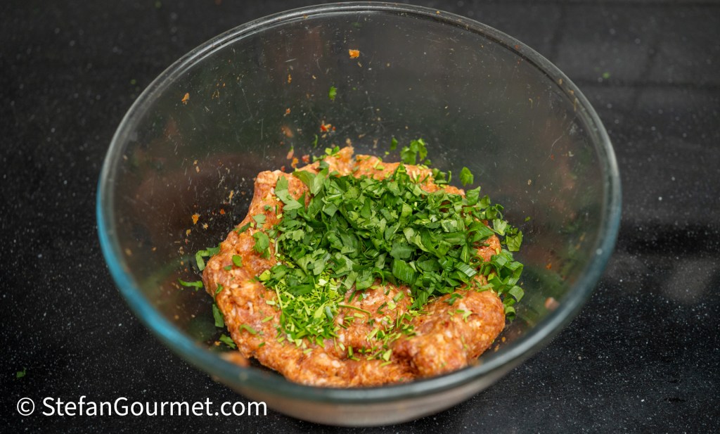 A glass bowl filled with a mixture of ground meat and finely chopped herbs, including cilantro, resting on a dark countertop.