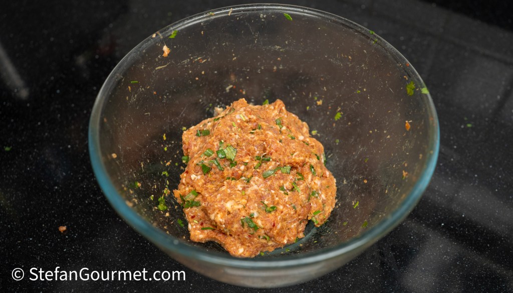 A glass bowl containing a mixture of ground meat, herbs, and spices, sitting on a dark countertop.