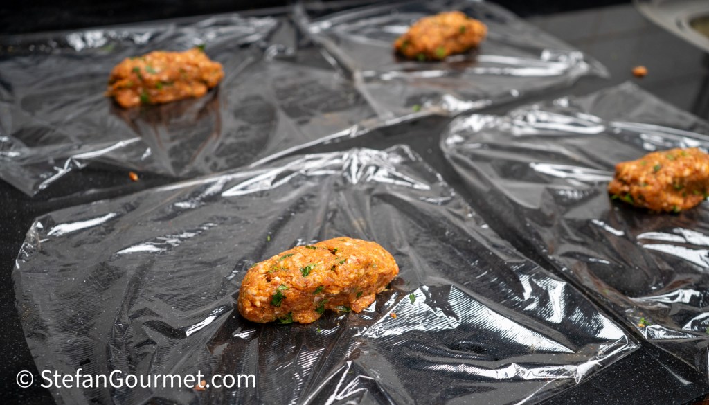 Five portions of meat mixture shaped into logs, resting on plastic wrap on a countertop.