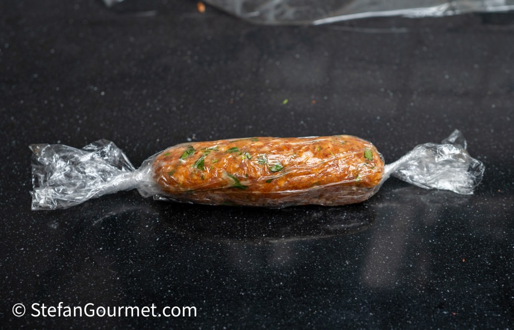 A rolled and wrapped food item in plastic wrap, displaying a mixture of ingredients with visible herbs, placed on a dark countertop.
