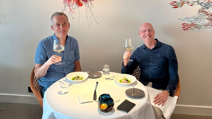 Two men smiling and toasting with wine glasses at a dining table set with white tablecloths and gourmet dishes in front of them.