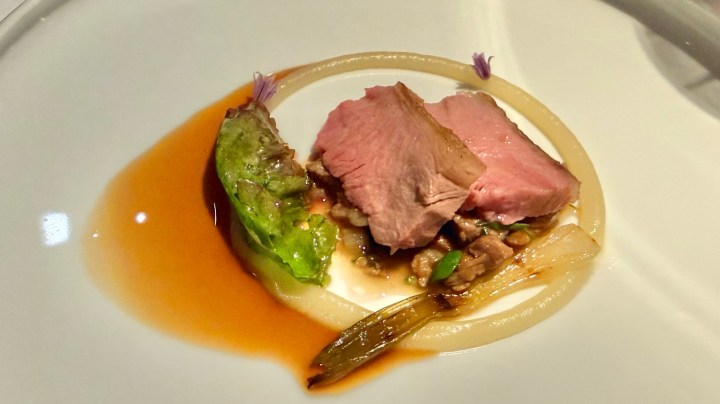 A gourmet dish featuring sliced pink meat, accompanied by greens and garnished with small flowers, served on a round white plate with a sauce around the edge.