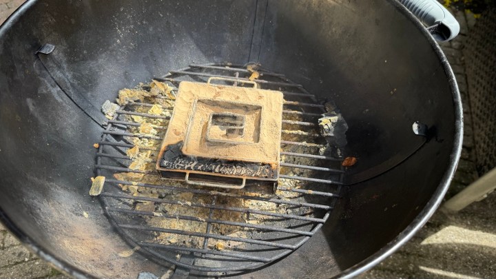 A close-up view of a grill with a square grill pan sitting atop it, surrounded by ash and remnants of previous cooking.