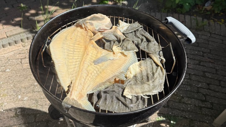 A black charcoal grill with a variety of fish being cooked, including a large fillet and smaller fish pieces on the grill rack.