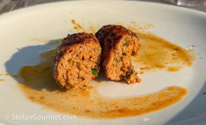 Two pieces of grilled sausage cut in half, revealing a moist and textured interior with herbs and spices on a white plate, accompanied by a light sauce.