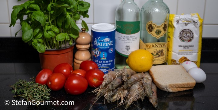 An assortment of ingredients for cooking, including fresh basil, tomatoes, shrimp, a lemon, bread, eggs, and various bottles of oil and salt arranged on a black countertop.