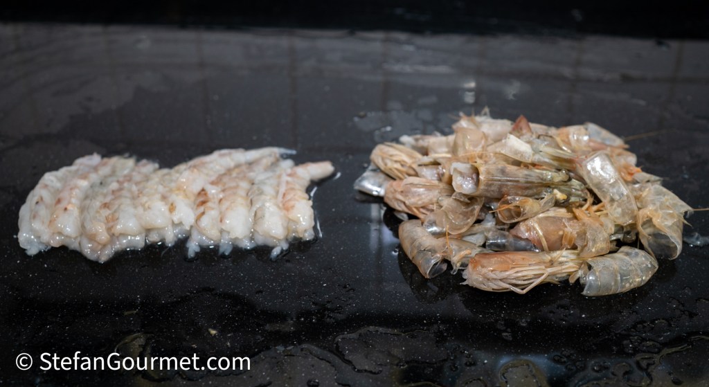 Raw shrimp tails on a black surface, with a separate pile of whole shrimp beside them.