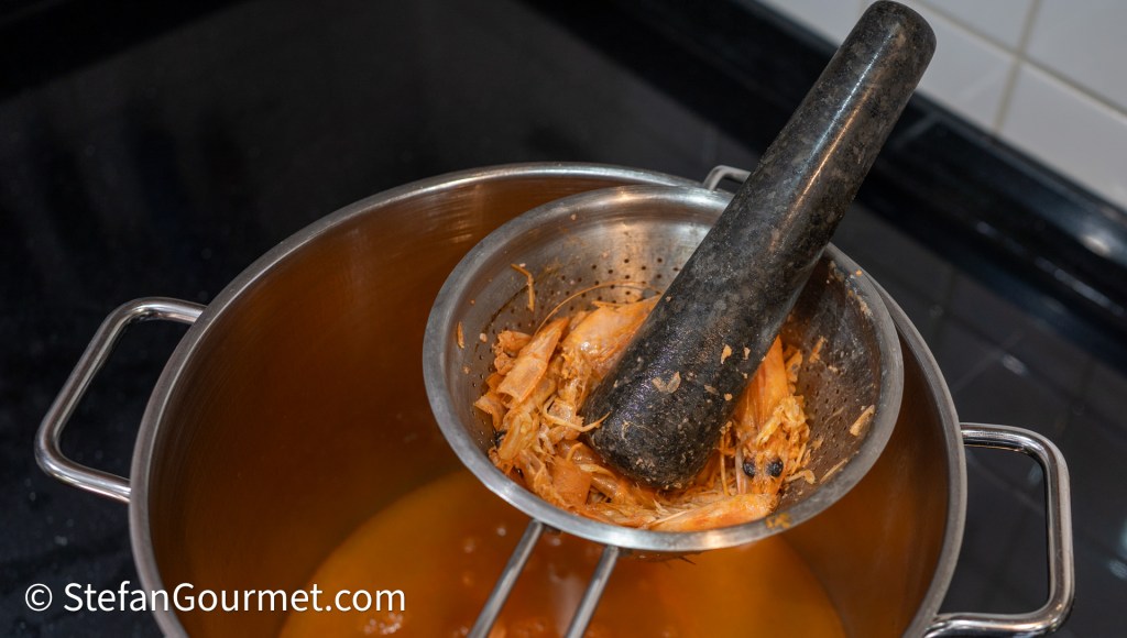 A pot of broth with a strainer containing shrimp shells and a pestle resting on top.