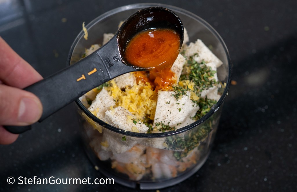 A person is adding a spoonful of sauce to a food processor containing pieces of bread, herbs, and orange zest.