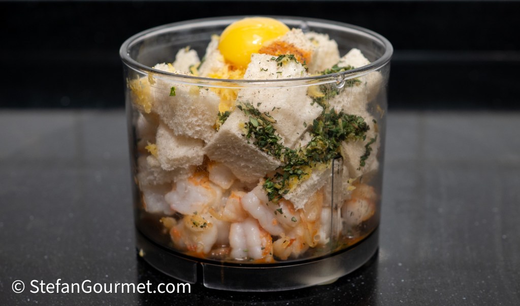A glass bowl containing shrimp, bread pieces, an egg yolk, and herbs, prepared for mixing.