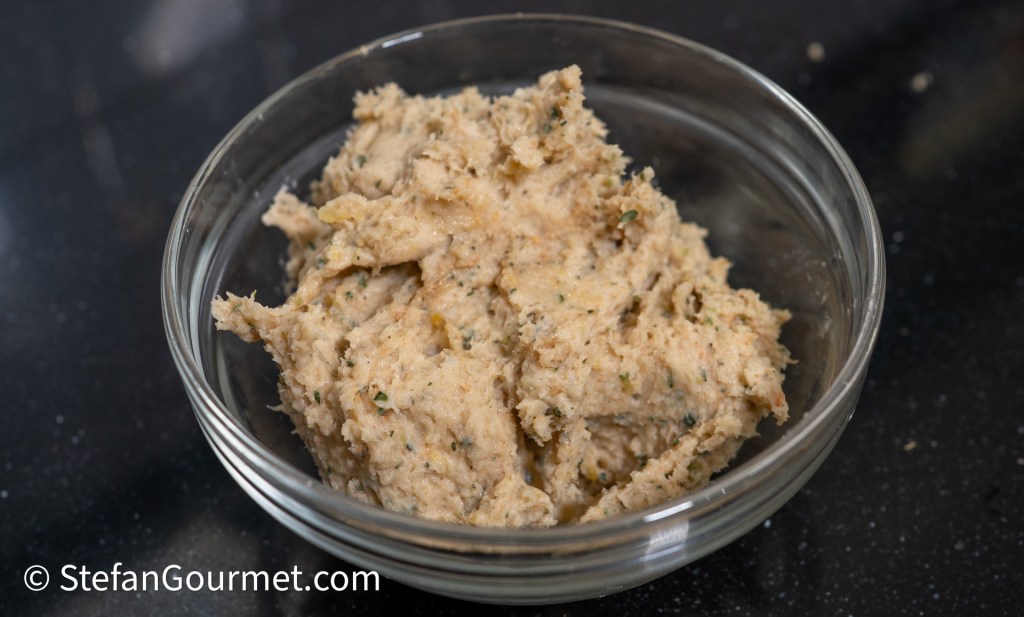 A bowl of creamy, textured spread made from blended ingredients, featuring herbs and spices, placed on a dark countertop.