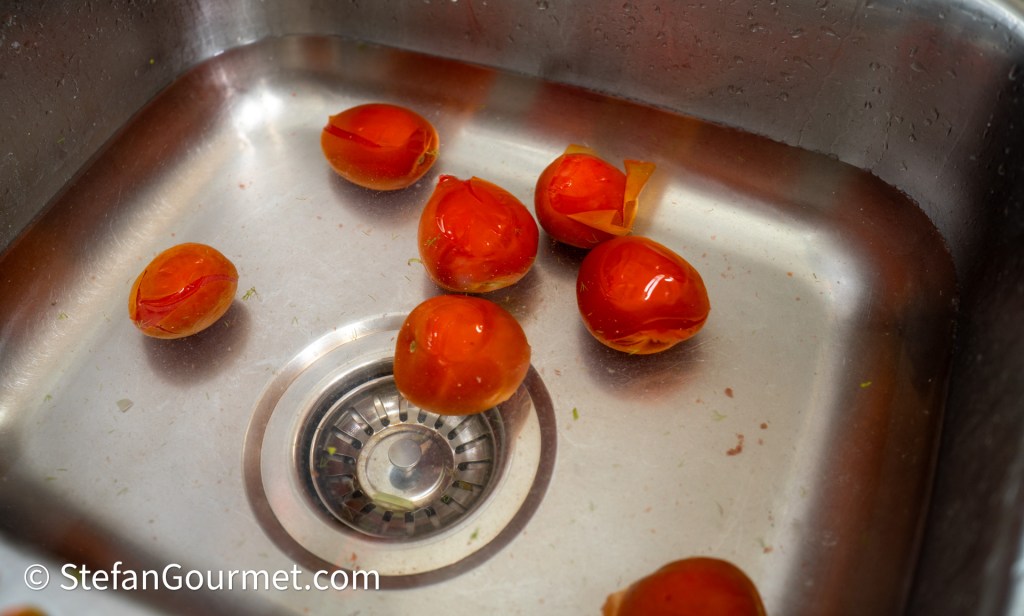 Red tomatoes with partially removed skins floating in a stainless steel sink.