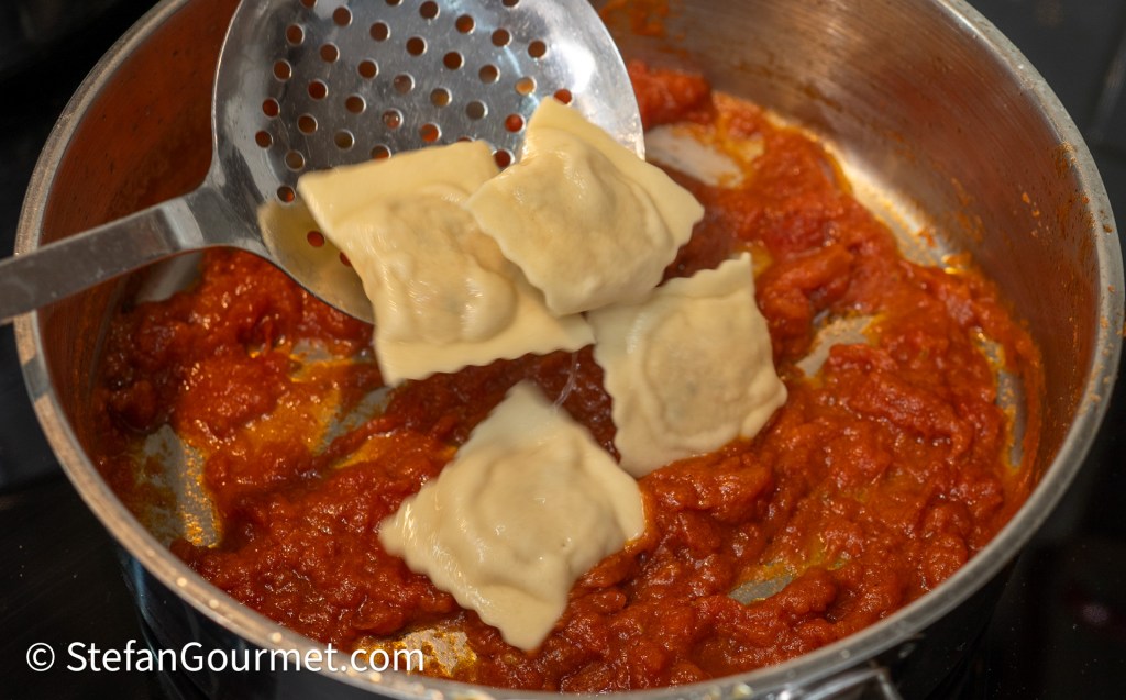 A slotted spoon lifting freshly cooked ravioli and placing them into a pot of simmering tomato sauce.