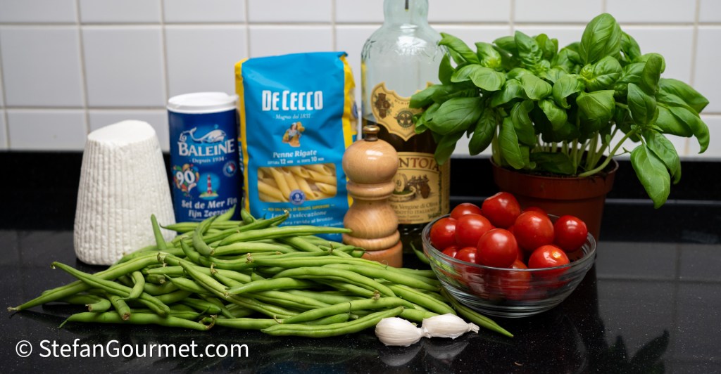 A collection of fresh ingredients for cooking, including green beans, cherry tomatoes in a glass bowl, a pot of fresh basil, a bottle of olive oil, a packet of penne rigate pasta, a container of sea salt, a pepper mill, and two cloves of garlic, displayed on a black countertop.